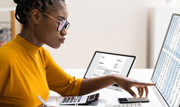 A woman works at a desktop computer, she has a tablet device and calculator on the desk beside her.