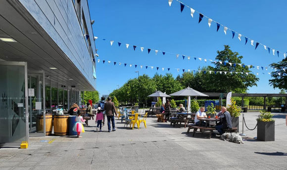 People sitting at picnic tables on a sunny day with blue skies outside Queen Margaret University's Students' Union building